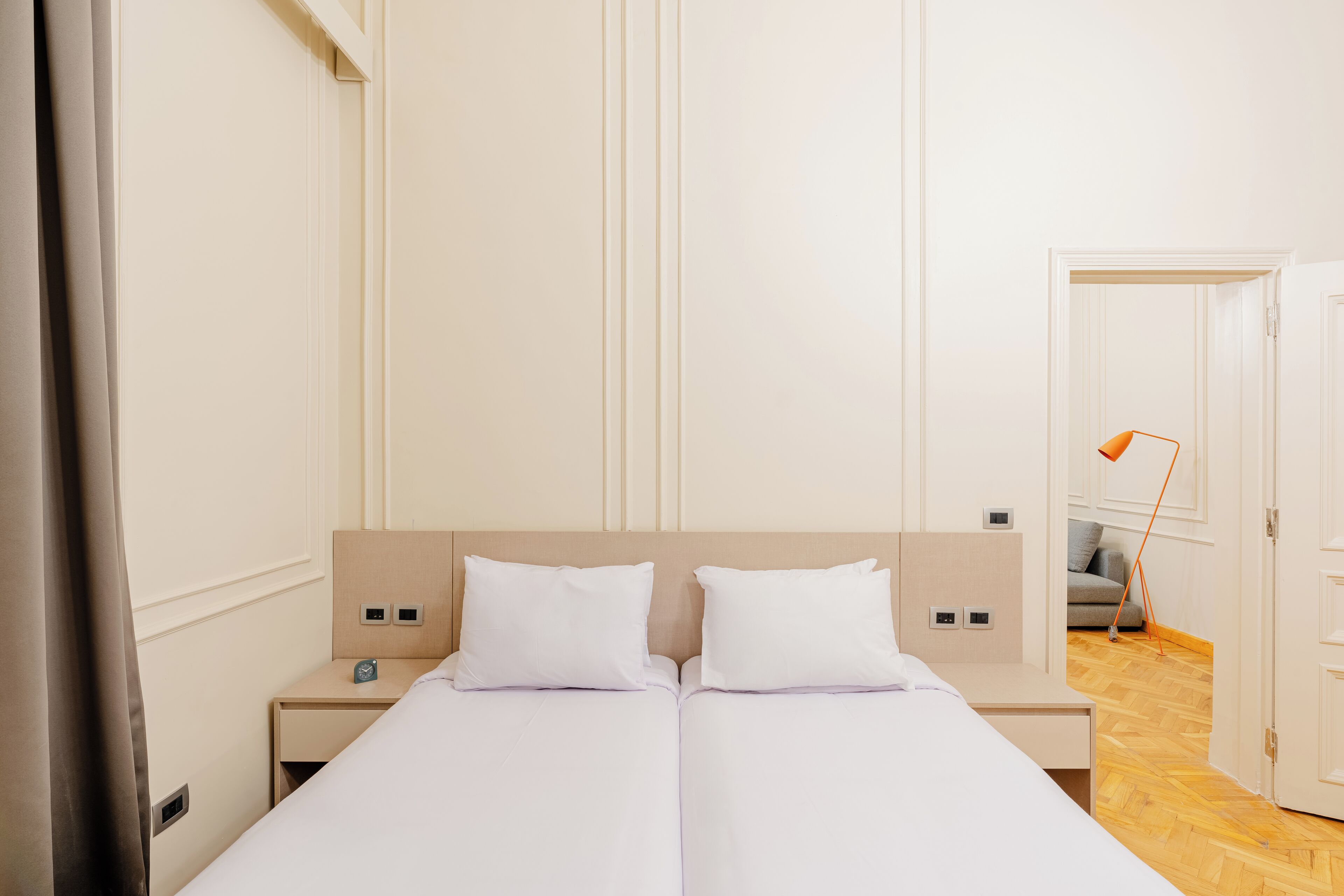 Una cama doble con ropa de cama blanca, dos mesitas de noche, una lámpara y una puerta que da a otra habitación.