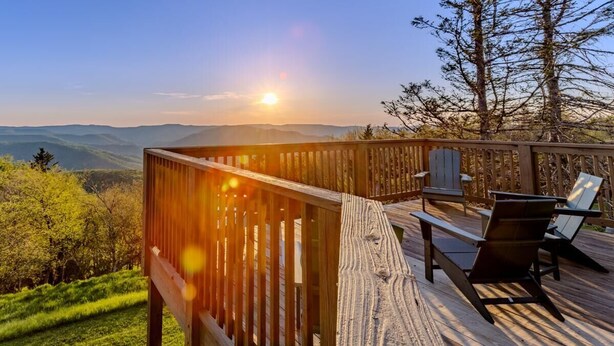 Una terraza de madera con vistas a montañas y un atardecer. Hay dos sillas y una mesa en la terraza.