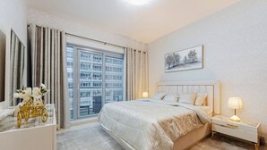 Ein Schlafzimmer mit einem großen Bett, einem Nachttisch, einer Kommode, einem Fenster mit Vorhängen und Blick auf die Stadt.