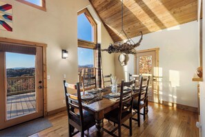A dining area with a wooden table set for four, large windows, and a view of the outdoors. - Some Place Else Cottage (Murphy)