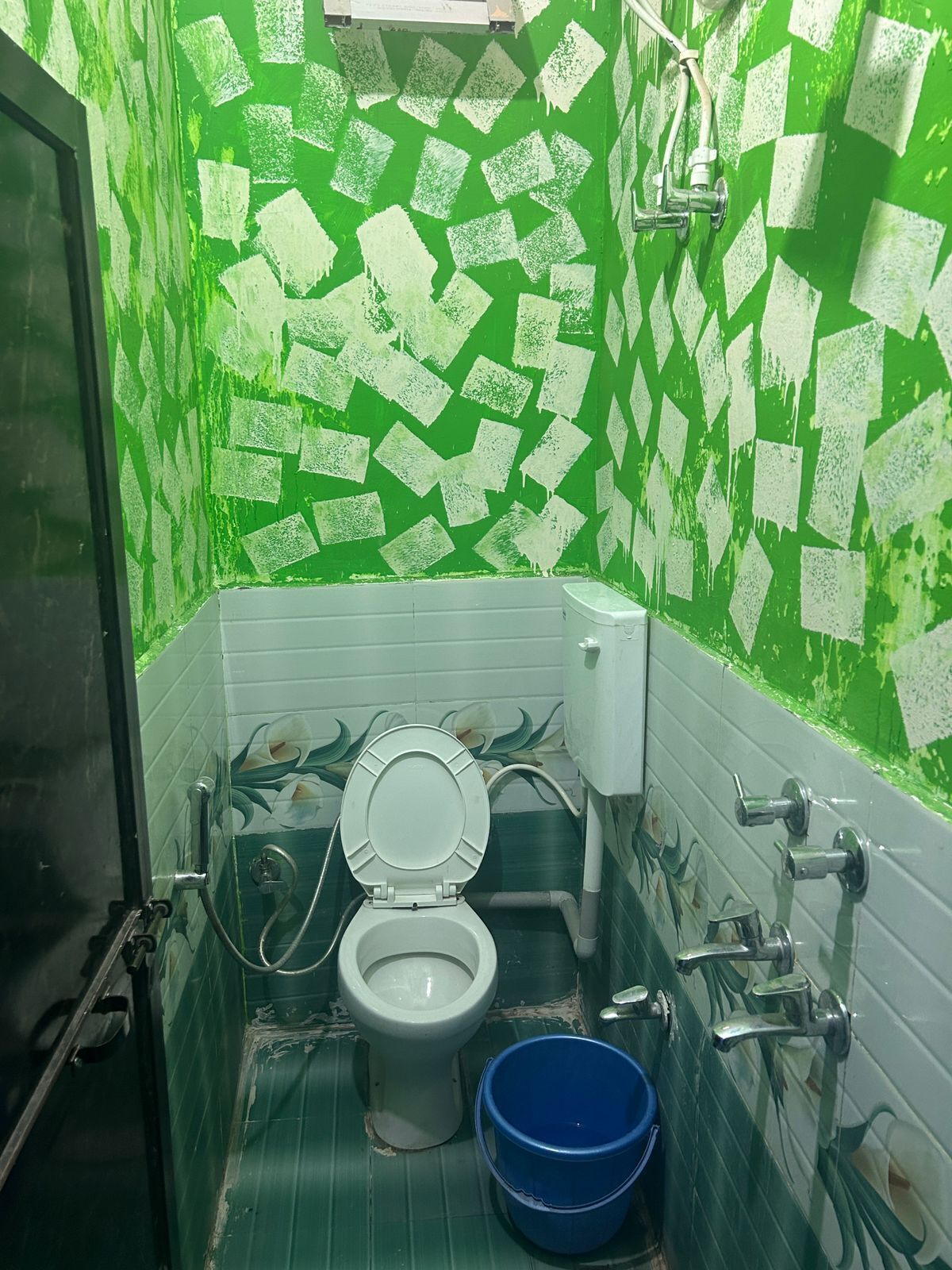 Une salle de bain avec un carrelage à motifs à carreaux verts et blancs, des toilettes, un seau bleu et une douche.