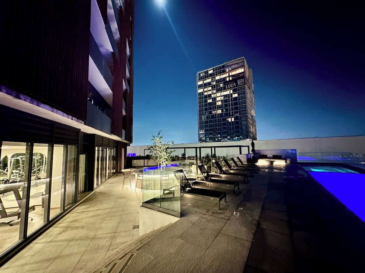 Una terrassa a la teulada amb seients exteriors, una piscina i vistes a un edifici alt al capvespre.