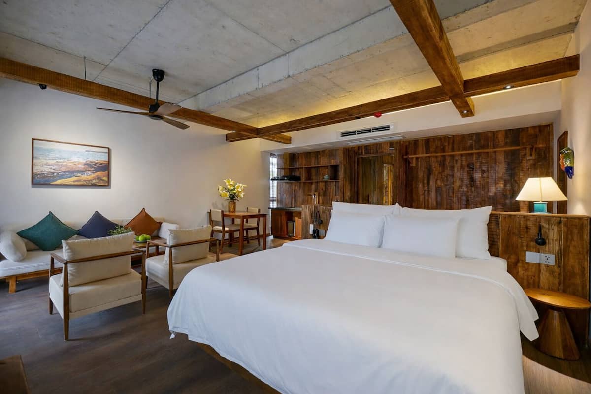 A bedroom with a large bed, wooden ceiling beams, a dining area with a table and chairs, and a wall-mounted shelf with decorative items.