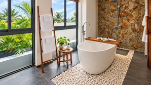Ein modernes Badezimmer mit freistehender Badewanne, einer Duschwand aus Stein und Blick auf Palmen durch große Fenster.