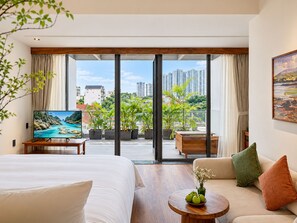 A hotel room with a bed, a sofa, a TV, a balcony with plants, and a city view. - Nexus House Retreat Xuan Thuy (Ho Chi Minh City)
