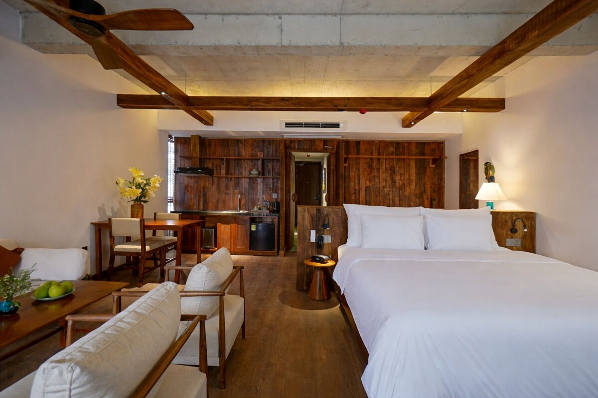 A hotel room with a wooden ceiling, a bed, a desk with chairs, a small kitchenette, and a dining area.