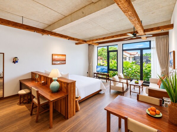 A hotel room with a bed, desk, and seating area, large windows with a view of greenery and buildings, and wooden beams on the ceiling.