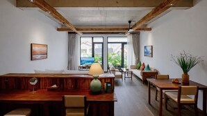 A spacious living area with a wooden dining table, a sofa, and a coffee table. There are two windows with curtains, a painting on the wall, and a plant on the right side. - Nexus House Retreat Xuan Thuy (Ho Chi Minh City)