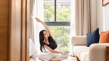 A person sitting on a yoga mat in a room with a large window, a sofa, and a painting on the wall.