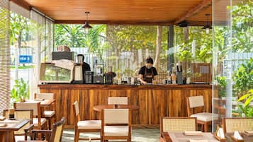 A cafe with wooden furniture, a barista preparing drinks, and large windows with greenery outside.