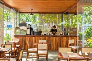 Un café avec des meubles en bois, un barista préparant des boissons et de grandes fenêtres donnant sur un espace verdoyant.