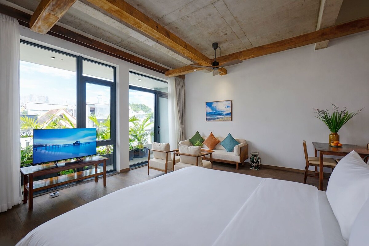 A hotel room with a large bed, a TV on a wooden stand, a ceiling fan, and large windows offering a view of palm trees and buildings.