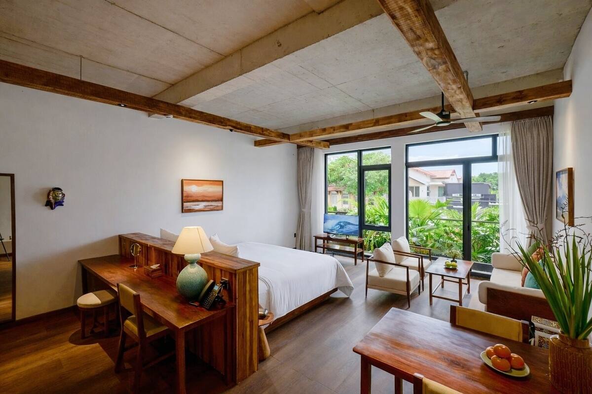 A bedroom with a bed, desk, and seating area, large windows with a view of greenery, and wooden beams on the ceiling.