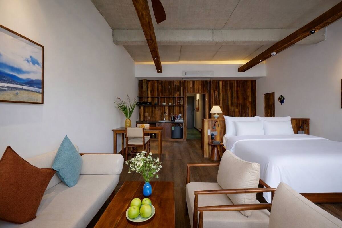 A hotel room with a bed, a sofa, a dining table with apples and flowers, a wooden wall, and a kitchen area.