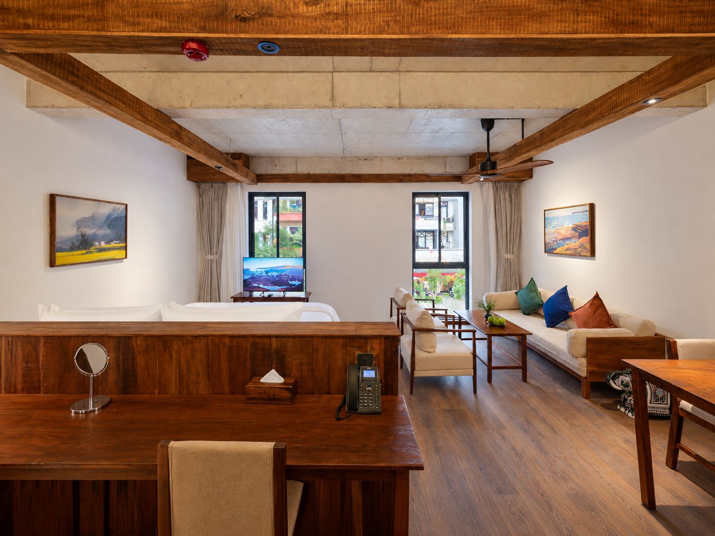 A hotel room with a wooden ceiling, a bed, a sofa, a dining table, and two windows offering views of the outside.