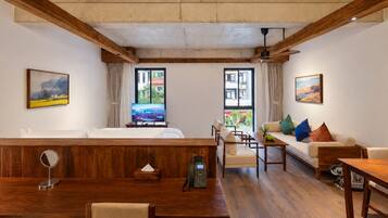 A hotel room with a wooden ceiling, a bed, a sofa, a dining table, and two windows offering views of the outside.