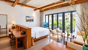 A hotel room with a bed, a desk, a sofa, and a dining table. There is a painting on the wall and a view of the outside through large windows.