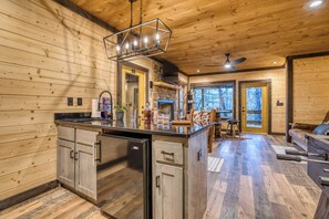 A wooden cabin interior with a kitchen island, a stone fireplace, and a dining area with a view of the forest. - Swanky Fox - hot tub-modern-fireplaces-pets-games (Cherry Log)