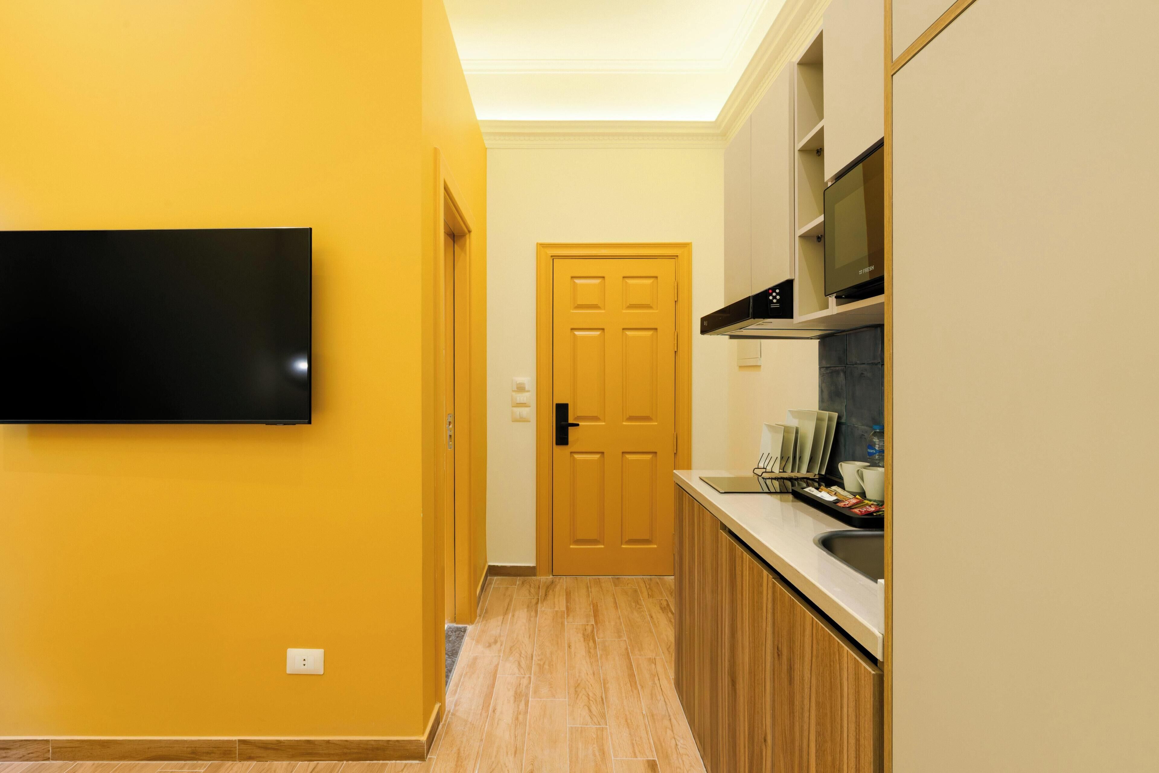 A hallway with a wooden door, a flat-screen TV mounted on the wall, and a kitchen area with cabinets and a countertop.