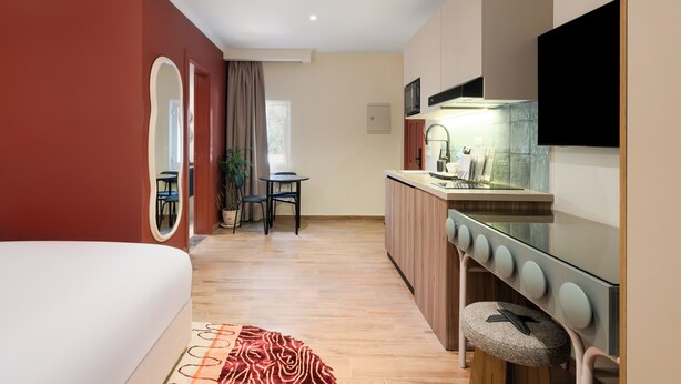 Ein kompaktes Hotelzimmer mit Bett, einer kleinen Küchenzeile, einem Essbereich mit Tisch und Stühlen sowie einem großen Spiegel.