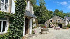 A stone building with white shutters and a covered entrance, surrounded by greenery and a gravel courtyard with outdoor seating. - Manoir du Menez Bihan - The refurbished gite - Site overlooking the Bay of Audierne (Audierne)