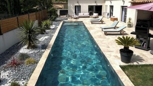 Una piscina rectangular con agua cristalina, rodeada por una solana empavedada. Se observan sillas de descanso y una mesita en un lado, y una zona cubierta con una parrilla en el otro.