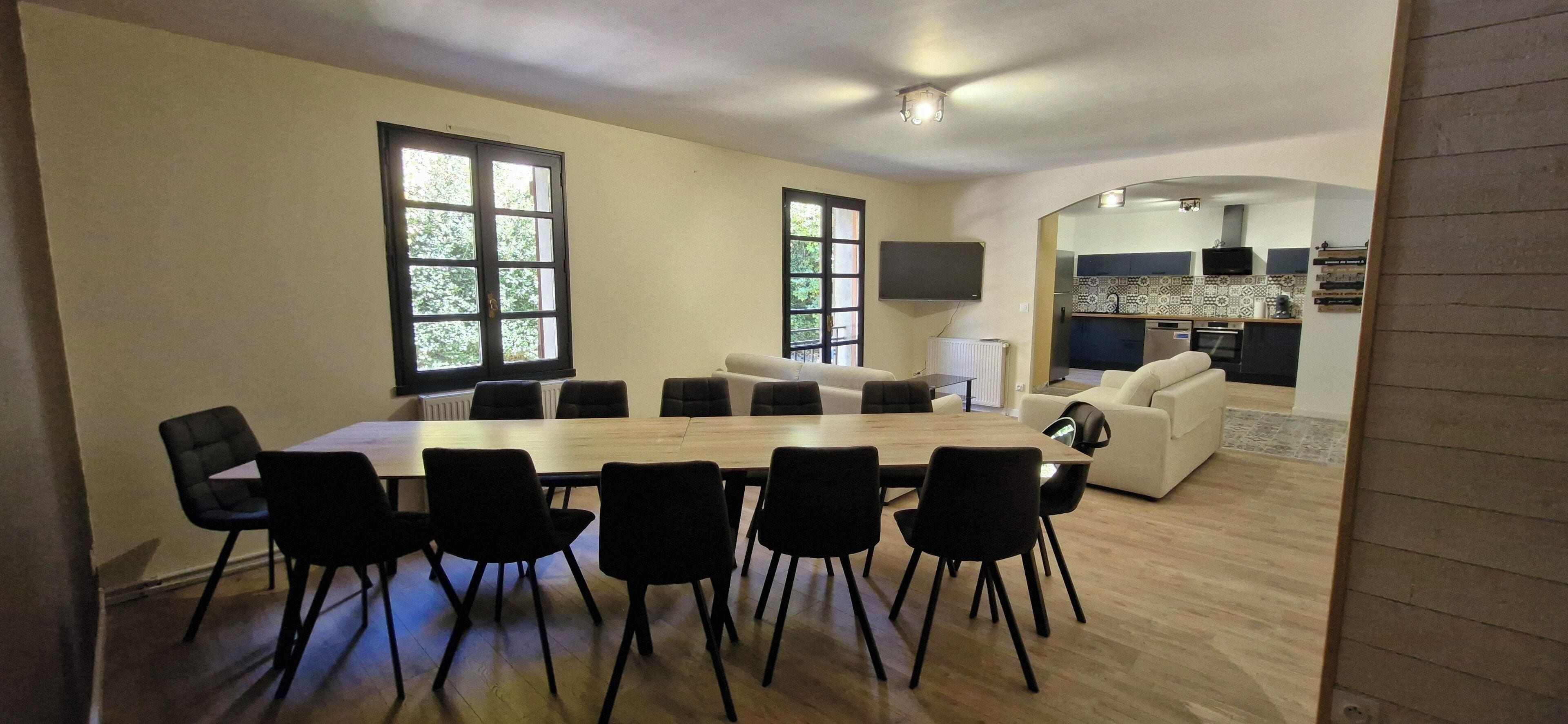A spacious living area with a large dining table surrounded by chairs, a sofa, and a kitchen area visible through an archway.