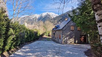 Une allée de gravier mène à un bâtiment doté d’un toit en pente, entouré d’arbres et avec des montagnes en arrière-plan.