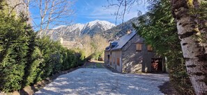 Une allée de gravier mène à un bâtiment doté d’un toit en pente, entouré d’arbres et avec des montagnes en arrière-plan.