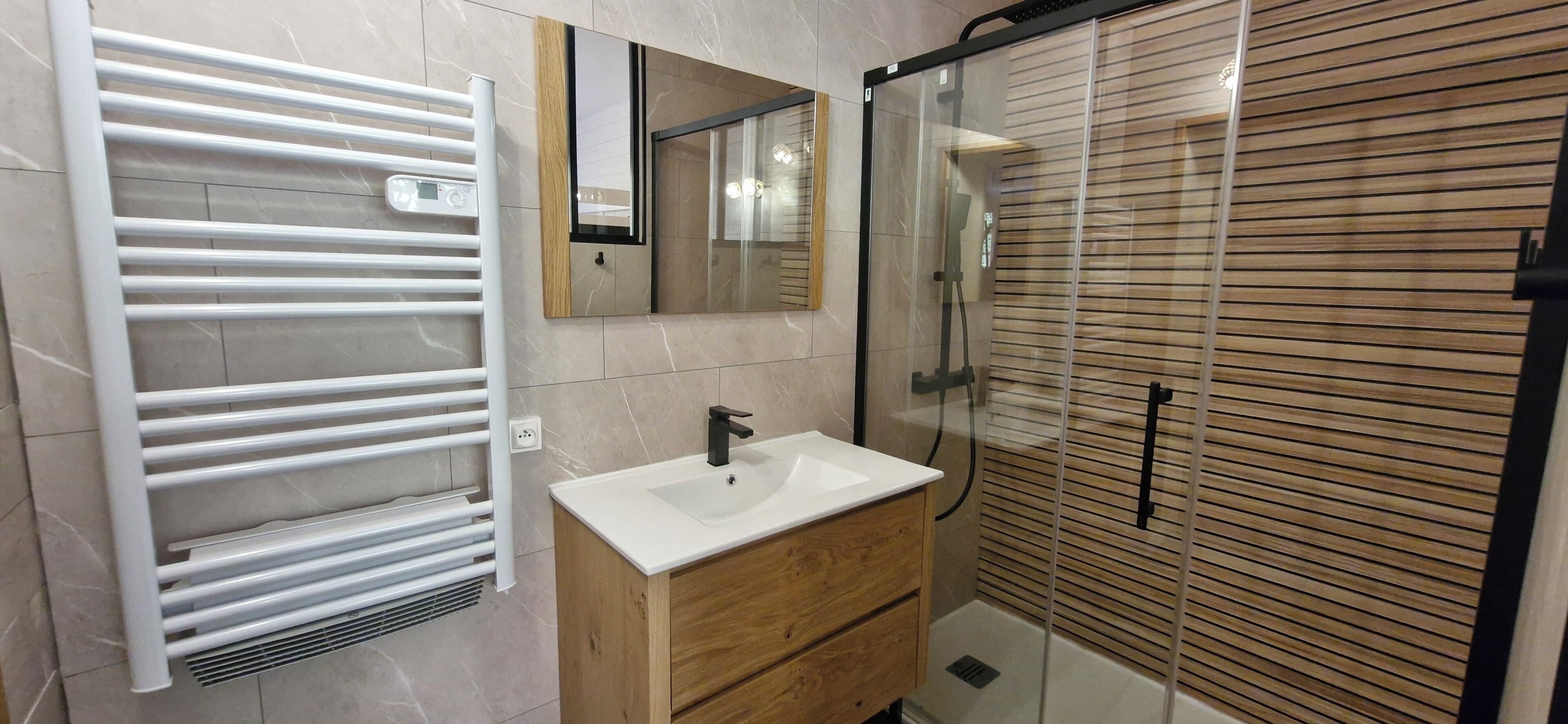 Een moderne badkamer met een witte handdoekhouder, een houten wastafelmeubel met een wastafel, een spiegel en een douche met glazen wanden en houten luiken.
