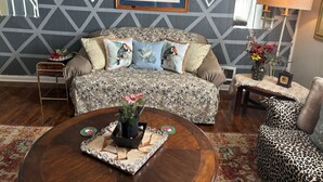 Uma sala de estar com um sofá estampado, uma mesa de centro redonda com um tabuleiro decorativo, uma poltrona com estampa de leopardo e uma parede com padrão.