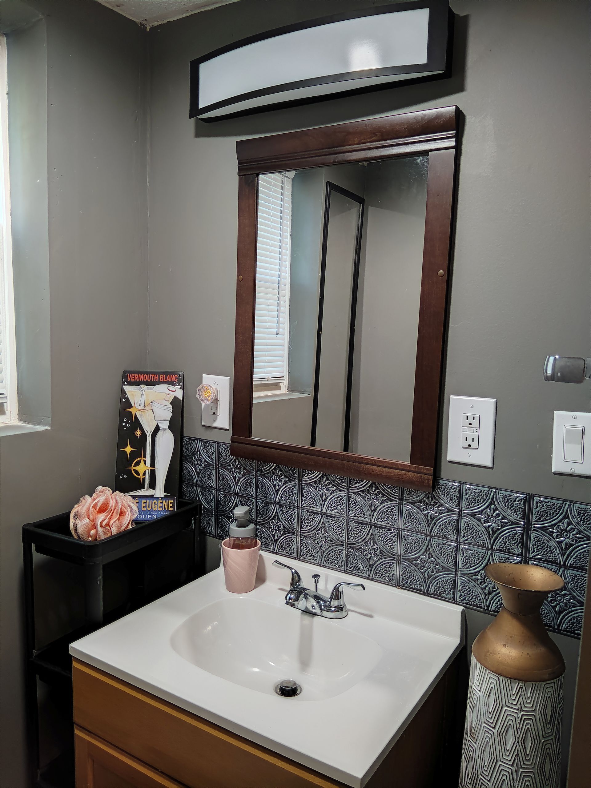 Une salle de bain avec un lavabo blanc, un miroir avec un cadre en bois, un vase décoratif et un mur recouvert de carreaux.