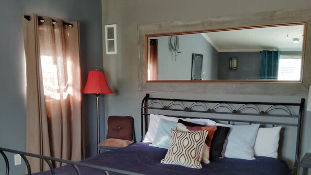 Um quarto com estrutura de cama em metal, um espelho grande acima da cama, um abajur vermelho e um quadro na parede.