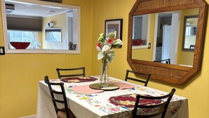 Uma área de jantar com parede amarela, um espelho grande e uma mesa posta com toalha de mesa florida e cadeiras.
