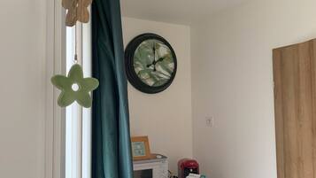 A room with a window, a curtain, a wall clock, a small table with a basket, and a wooden door.