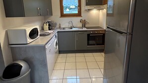 A compact kitchen with grey cabinets, a stainless steel refrigerator, a microwave, a sink, and a window above the sink. - Cosy chalet 2-8pers/linen provided (Gérardmer)