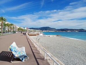 Eine Uferpromenade mit Kiesstrand, Bänken und Blick auf das Meer und die entfernten Hügel.