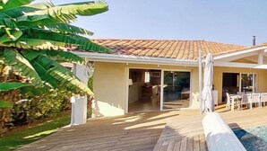A wooden deck leading to a house with a tiled roof, a pool, and a patio area with outdoor furniture.