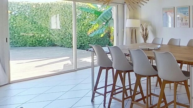 A dining area with a wooden table and chairs, large windows, and a ceiling fan.