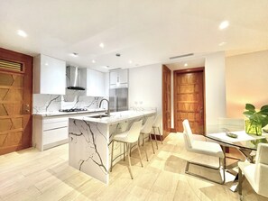 A modern kitchen with white cabinetry, a marble island, stainless steel appliances, and a dining area with white chairs and a glass table. - Lovely luxury 2-bedroom 3 beds apartment with AC in Casa de Campo (Casa de Campo)