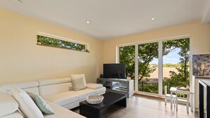 Una sala de estar moderna con un sofá, una mesa de centro y un televisor. Hay una puerta corrediza de vidrio que da a un balcón con vistas a zonas verdes y un paisaje distante.