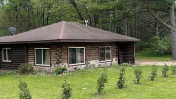 A log cabin with a sloped roof, surrounded by a grassy yard and small trees.