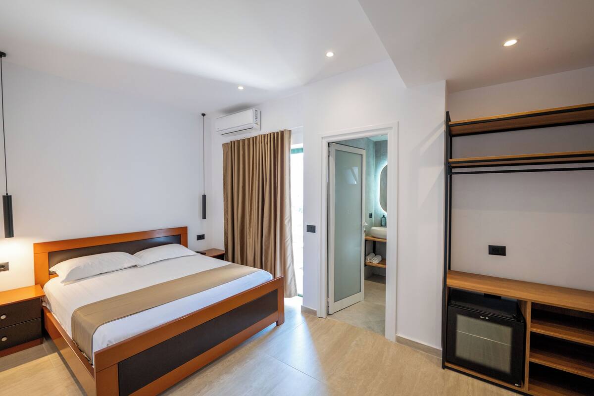 Habitació d'hotel moderna amb un llit de fusta, un escriptori integrat, una petita cuina americana i una porta corredissa de vidre que condueix a un bany.