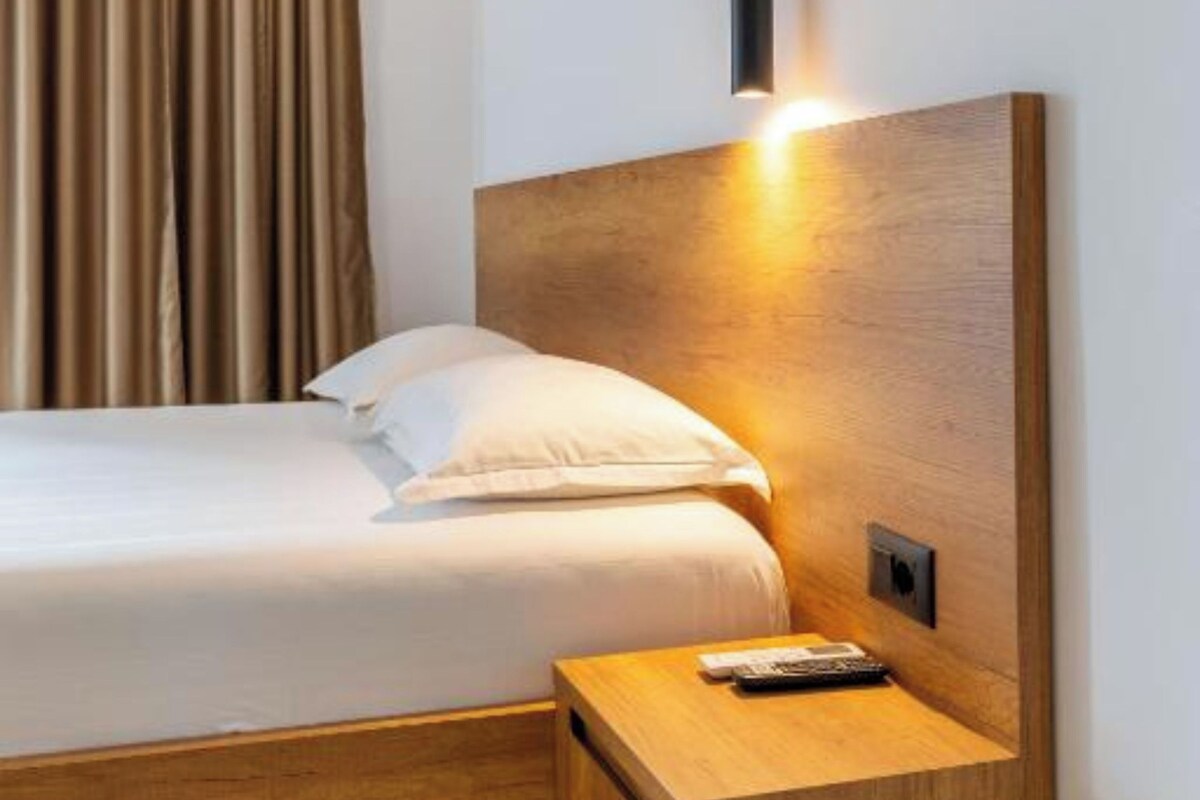 A modern hotel room with a wooden headboard, a bedside table with a remote control, and a curtain with a warm light behind it.