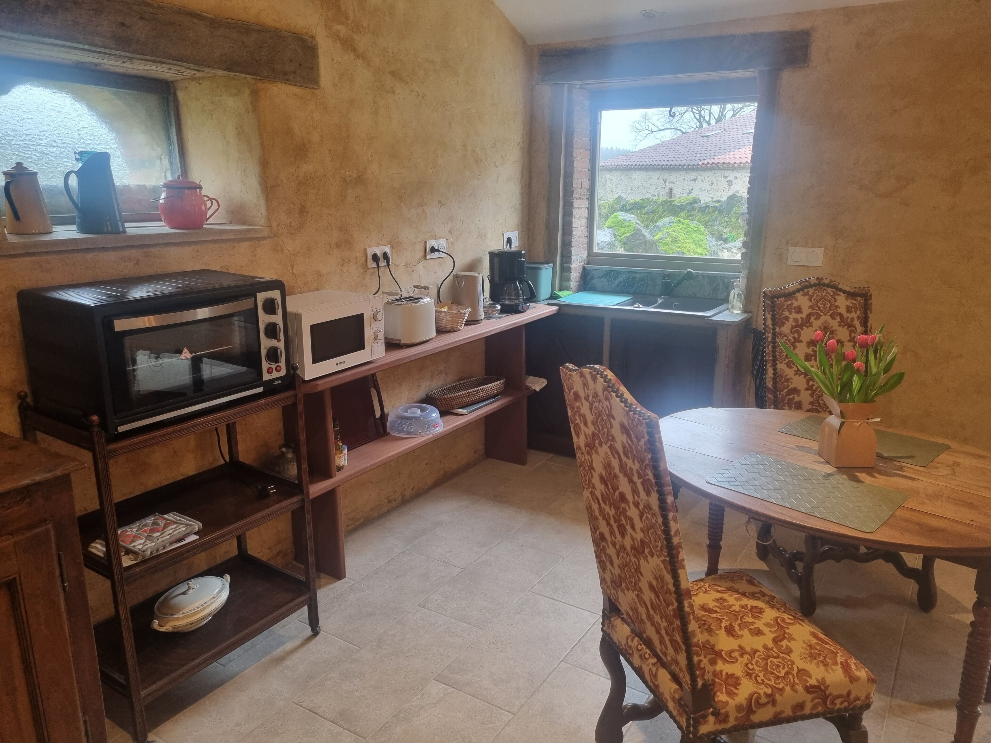 Eine Küche mit Mikrowelle, Toaster und Kaffeemaschine auf der Arbeitsplatte, einem Esstisch mit Stuhl und einem Fenster mit Blick auf Grünflächen.