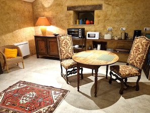A cozy dining area with a round wooden table, two patterned chairs, a wicker chair with a yellow cushion, a small cabinet with kitchen appliances, and a decorative rug.