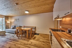A cozy room with a wooden ceiling, a dining table with chairs, a sofa, and a kitchen area with wooden cabinets and a sink. - Appartement Omesberg 1 (Lech am Arlberg)