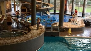 An indoor swimming pool with multiple levels and slides, surrounded by large windows.