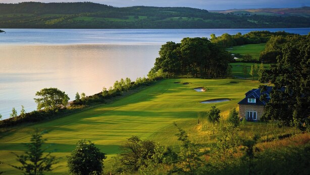 A golf course with a clubhouse, surrounded by trees and hills, overlooking a body of water.