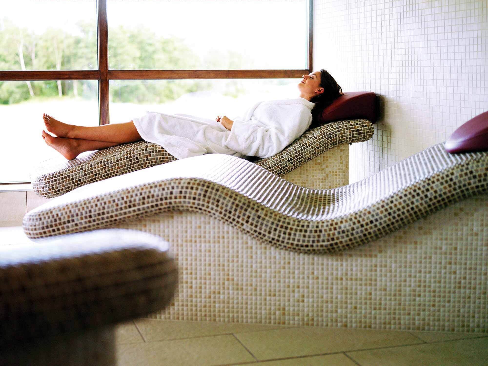 A person lying on a textured lounge chair in a room with a tiled wall and a window.